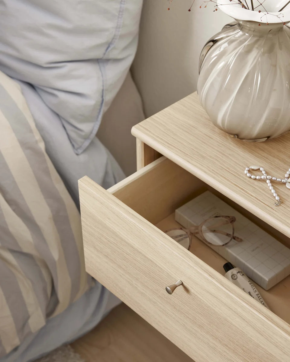 A light natural wood nightstand with an open drawer, styled next to a bed with gray and white striped bedding. A ribbed white ceramic vase with dried branches and a pearl bow sit on the nightstand.