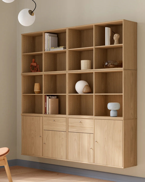 A large light oak wood wall-mounted bookcase with open shelves on the upper portion and three cabinet doors with two center drawers below, styled with books and decorative objects in a room setting.