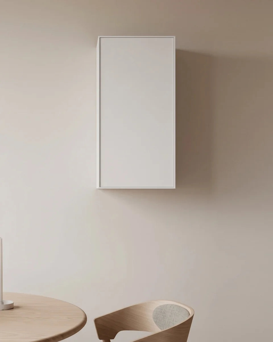A minimalist white rectangular wall-mounted cabinet or storage box shown in a clean interior setting with a beige wall, partial wooden table, and chair.