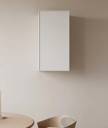 A minimalist white rectangular wall-mounted cabinet or storage box shown in a clean interior setting with a beige wall, partial wooden table, and chair.